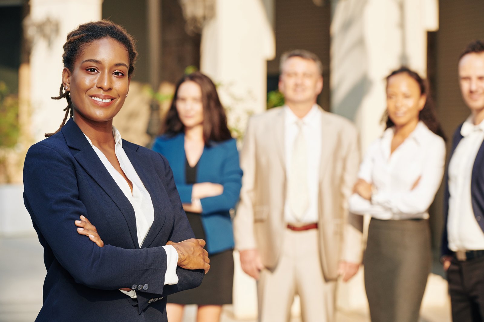 XTRM Group | Beautiful confident successful businesswoman standing with arms folded and smiling at camera, her colleagues standing in background
